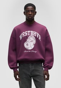 Felpa viola con scollo rotondo, vestibilità oversize e testo grafico bianco. Gli accenti includono un design stampato con un piccolo volto.