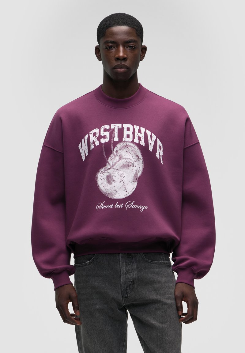 Lilla sweatshirt med rund halsudskæring, oversized pasform og hvid grafisk tekst. Detaljerne inkluderer et trykt design med en lille ansigt.