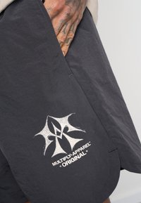 Pantaloni corti neri realizzati in tessuto leggero con orlo curvo. Logo bianco con la scritta "MULTIPLY APPAREL ORIGINAL" sul lato.