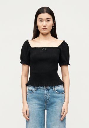 Žena s dlhými tmavými vlasmi má na sebe čierny top so štvorcovým výstrihom a nadýchanými rukávmi a modré džínsy, stojí pred jednofarebným svetlým pozadím.