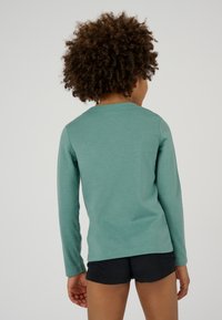 Camicia verde a maniche lunghe realizzata in un tessuto morbido, con scollatura rotonda e maniche aderenti. Indossata con pantaloni corti neri, vista da dietro.