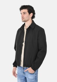 Giacca nera con zip realizzata in tessuto leggero, presenta un colletto, polsini elasticizzati e un taglio casual. Indossata su una camicia beige.