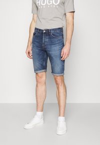 Vaaleansiniset farkkushortsit, joissa on käännetty helma ja klassinen viisi taskua, yhdistettynä harmaaseen logopaitaan ja valkoisiin lenkkareihin.