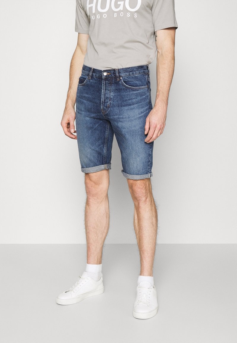 Vaaleansiniset farkkushortsit, joissa on käännetty helma ja klassinen viisi taskua, yhdistettynä harmaaseen logopaitaan ja valkoisiin lenkkareihin.