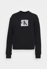 Sudadera negra con cuello redondo y mangas largas. Presenta un parche rectangular blanco con texto negro en superposición en la parte delantera.