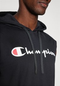 Schwarzer ärmelloser Hoodie mit einer vorderen Kängurutasche, der ein auffälliges weißes "Champion"-Logo und einen roten Akzent aufweist. Weiches Baumwollmaterial.
