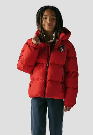 Rote Pufferjacke mit Reißverschluss, elastischen Bündchen und Kapuze. Mit Logo-Patch auf der linken Brust. Getragen über einem weißen Shirt und dunklen Hosen.