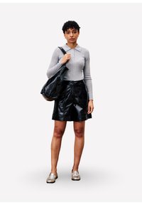 Vrouw met kort krullend haar, gekleed in een grijze lange mouw polo, zwarte leren shorts, zilveren instappers en houdt een grote zwarte schoudertas vast.