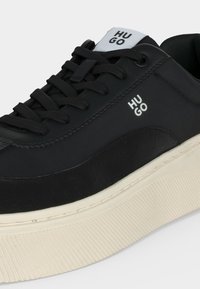 Μαύρο sneaker χαμηλού πάνω μέρους με παχιά υπόλευκη σόλα, μαύρα κορδόνια και λευκή επιγραφή "HUGO" στο πλάι και στη γλώσσα.