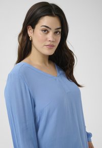Blouse légère bleu clair à manches longues avec un col en V et un tissu texturé. Présente une couture frontale discrète et une coupe décontractée.