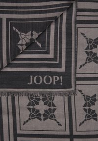 Geweven sjaal met een geometrisch patroon in zwart en grijs, met franjes aan de randen en een logo-label met de tekst "JOOP!" in een hoek.