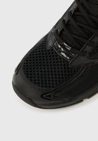 Pantof sportiv negru cu detalii din plasă, accente din piele texturată și vârf rotund. Dispune de șireturi și talpă de cauciuc pentru aderență.