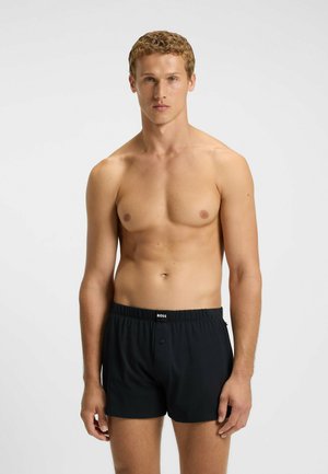 Junger Mann mit kurzem, lockigem Haar, der schwarze BOSS-Boxershorts trägt und vor einem schlichten, hellen Hintergrund mit neutralem Gesichtsausdruck nach vorn blickt.