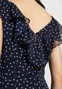 Navy blue dress with white polka dots, featuring a ruffled neckline and back zipper. The fabric appears lightweight and textured.