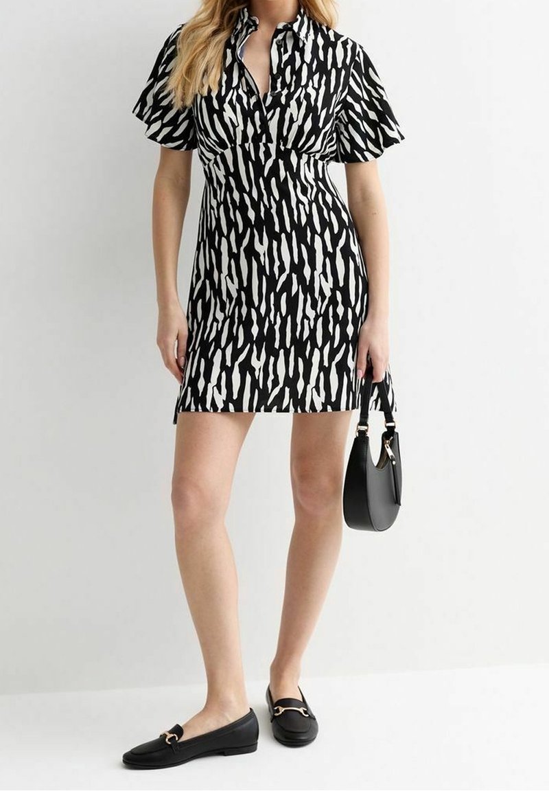 Černobíle vzorované šaty s límečkem a krátkými zvonovými rukávy, doplněné černými mokasínami a malou černou kabelkou.