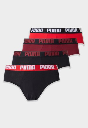 Muskularny mężczyzna stojący boso, ubrany w czarne slipy z czerwoną gumą z logo Puma na jednolitym białym tle.