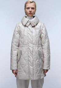 Cappotto imbottito di un colore crema chiaro con colletto alto, caratterizzato da una texture trapuntata, tasche laterali e chiusura frontale con hardware decorativo.