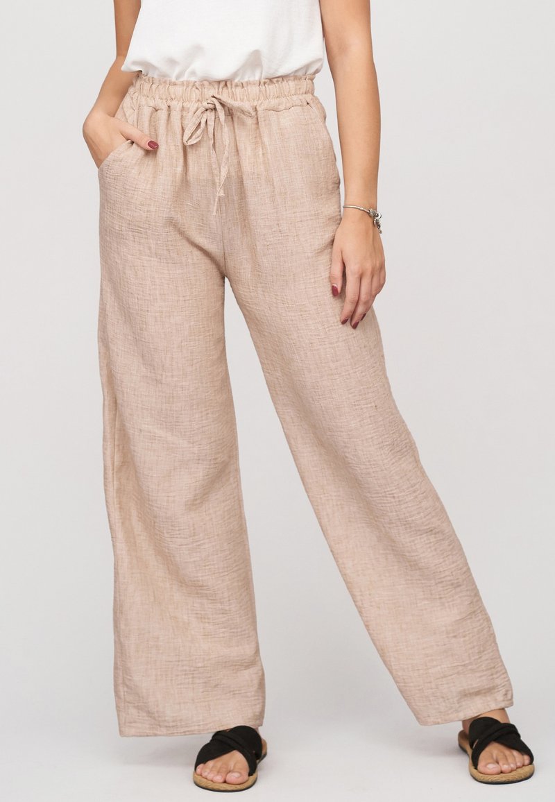 Femme portant un pantalon large en lin beige avec taille élastique et cordon, associé à des sandales slides noires et un top blanc.