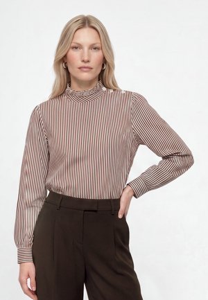 Kvinde med glat blondt hår bærer en langærmet brun og hvidstribet bluse og højtaljede mørkebrune bukser, stående med den ene hånd på hoften.