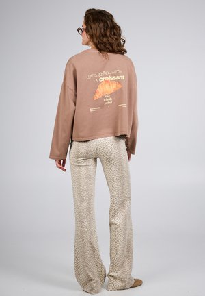 Donna con capelli ricci che indossa occhiali, una felpa marrone con grafica di croissant e testo, e pantaloni beige con stampa leopardata, in piedi contro uno sfondo grigio.