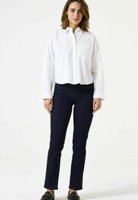 Chemise blanche courte boutonnée avec une poche avant, associée à un jean droit bleu foncé. Le mannequin porte des mocassins noirs.