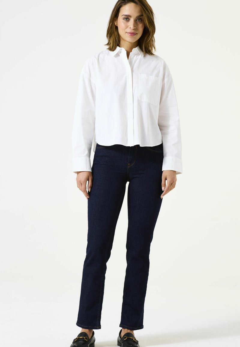 Chemise blanche courte boutonnée avec une poche avant, associée à un jean droit bleu foncé. Le mannequin porte des mocassins noirs.