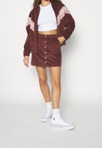 Vrouw in een bruine corduroy rok met knopen aan de voorkant en bijpassende jas met roze details, witte crop top, witte sokken en witte high-top sneakers.