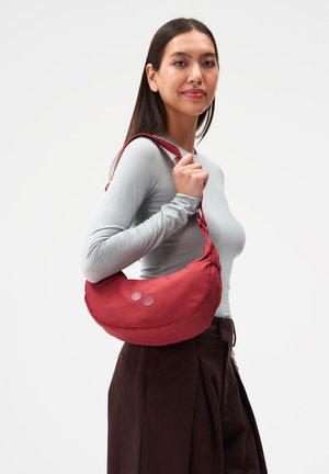 Junge Frau mit langen dunklen Haaren, die ein hellgraues langärmeliges Oberteil und dunkle Hosen trägt und eine rote, halbmondförmige Umhängetasche hält.