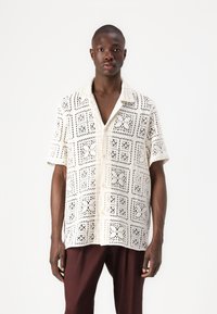 Chemise blanche à manches courtes en dentelle, ornée d'un motif de découpes géométriques et d'une fermeture par boutons, associée à un pantalon marron foncé.