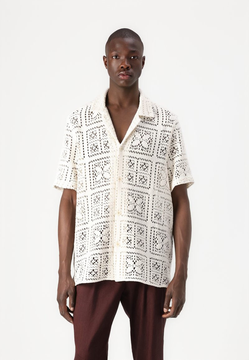 Chemise blanche à manches courtes en dentelle, ornée d'un motif de découpes géométriques et d'une fermeture par boutons, associée à un pantalon marron foncé.