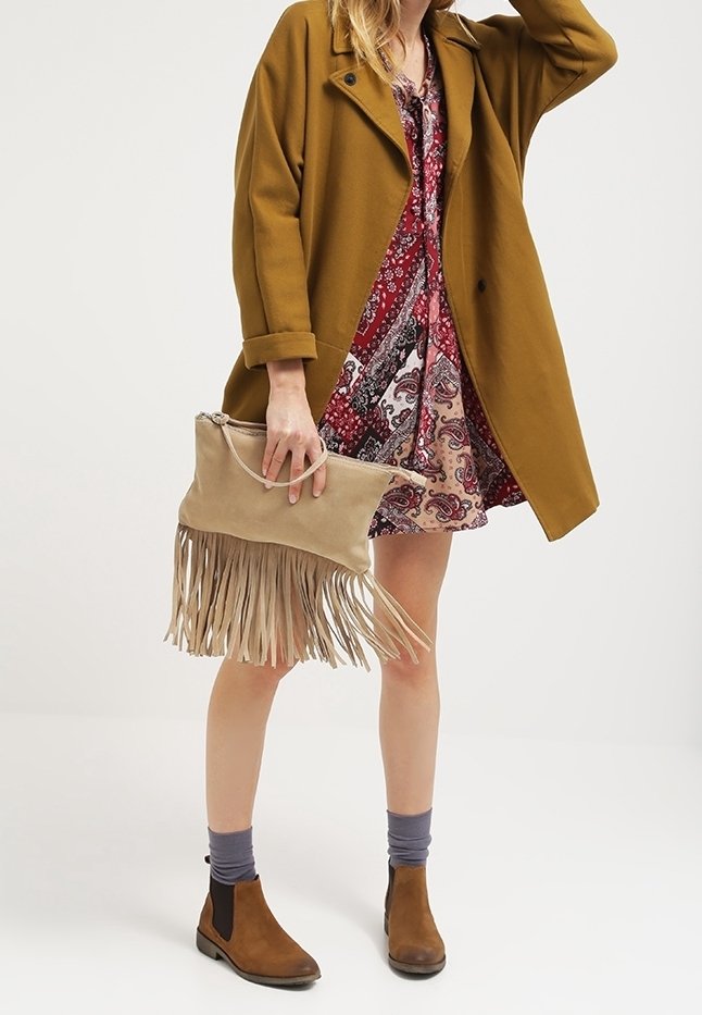 Femme portant un manteau moutarde sur une robe rouge à motifs, tenant une pochette beige à franges, portant des bottines marron et des chaussettes grises.
