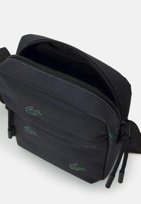 Borsa a tracolla nera con tasca anteriore con cerniera, loghi di coccodrillo ricamati in verde, tracolla regolabile e scomparto principale parzialmente aperto.