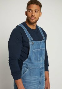 Salopette in denim con spalline regolabili, tasca frontale e tessuto leggero blu, indossata sopra una maglietta a maniche lunghe scura.