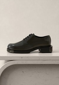 Μαύρο γυαλιστερό δερμάτινο παπούτσι για επίσημες περιστάσεις με κορδόνια μπροστά, τετράγωνα μεταλλικά στοιχεία στη μύτη και τετράγωνο τακούνι, ακουμπισμένο σε ανοιχτόχρωμη επιφάνεια.