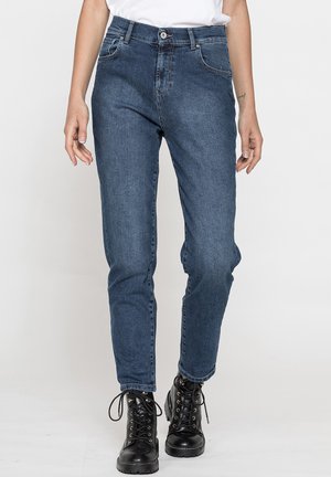 Donna che indossa jeans in denim blu e stivaletti neri con lacci, in piedi con le mani rilassate ai lati.