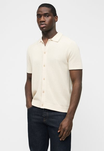 Άνδρας που φοράει κοντομάνικο, κρεμ πλεκτό πουκάμισο με κουμπιά και γιακά, συνδυασμένο με σκούρο μπλε τζιν, στέκεται με τα χέρια στις τσέπες.