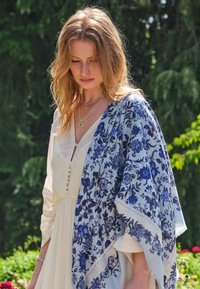 Witte jurk met knoopdetails, gecombineerd met een lichtblauwe sjaal met een bloemenpatroon in donkerblauw. Zachte stof, vloeiende silhouet.