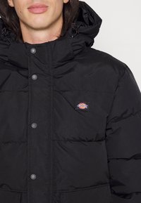 Giacca a vento nera imbottita con cappuccio, bottoni a pressione frontali, texture trapuntata e una piccola etichetta con logo sul lato sinistro del petto.