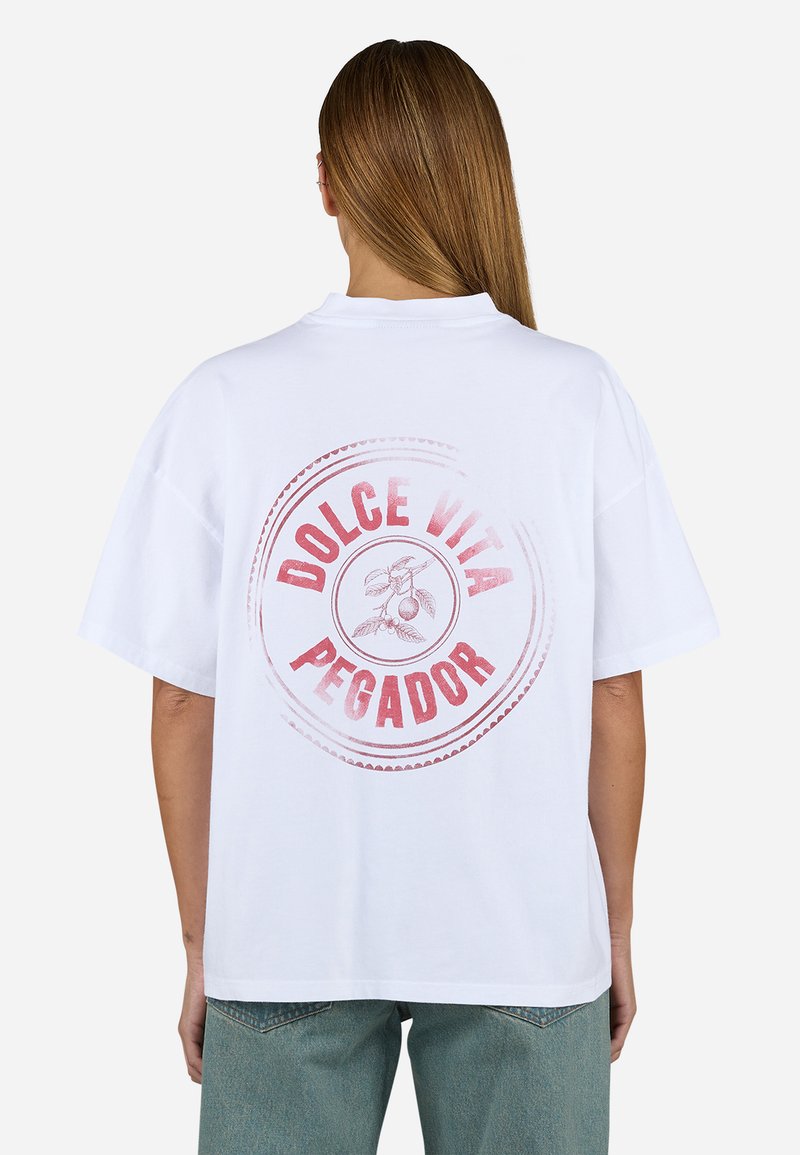 T-shirt oversized blanc présentant un imprimé circulaire rouge avec le texte "DOLCE VITA PEGADOR" et une illustration botanique. Manches courtes et coupe décontractée.