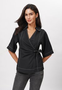Černá wrap top s krátkými rukávy, bílým kontrastním šitím, V-výstřihem a zavazováním v pase. Spárováno se šedými džíny. Hladká textura.