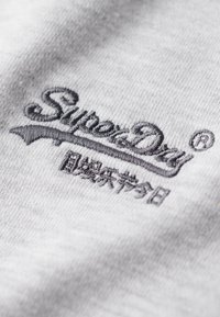 Tessuto grigio con il logo ricamato "Superdry" in nero, accompagnato da un accento decorativo sottostante e testo in un'altra lingua.
