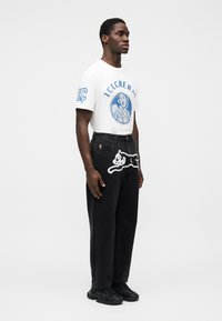 Λευκό μπλουζάκι με μπλε γραφικό και λογότυπο, συνδυασμένο με μαύρα τζιν με λευκό κεντητό σχέδιο. Μαύρα αθλητικά παπούτσια ολοκληρώνουν την εμφάνιση.