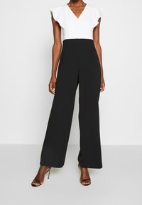 Svart och vit overall med V-ringning, korta volangärmar, vida ben och slät tygtextur.