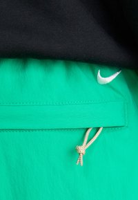 Shorts atletici verzi cu un brâu negru, având un mic logo alb Nike și un buzunar frontal cu închidere tip șnur incorporată.