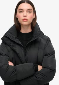 Svart puffjacka med hög krage, med dragkedjestängning och tryckknappar, samt en stickad svart tröja under. Slät textur.