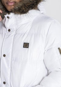 Biely pufferník s kapucňou obloženou umelou kožušinou, so zipsovým vreckom a malým kovovým logom na hrudi. Hladká, lesklá textúra látky.