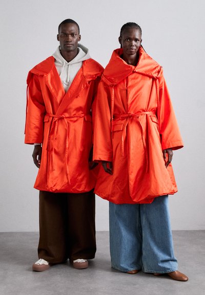 Δύο μοντέλα φορούν υπερμεγέθη, έντονα πορτοκαλί, σατινέ jackets με φαρδιές γιακάδες και ζώνες στη μέση, συνδυασμένα με φάρδους παντελόνια σε καφέ και τζιν.
