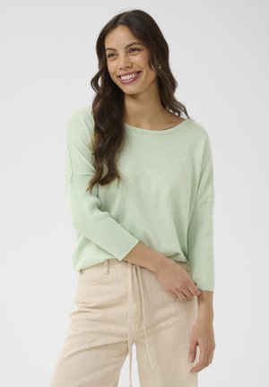 Usmievavá žena s dlhými tmavými vlasmi, oblečená v svetlozelenom tričku s dlhým rukávom a béžových nohaviciach so sťahovacou šnúrkou v páse, na jednoduchom pozadí.