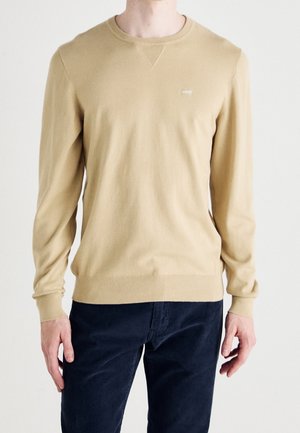 Homme portant un pull beige à manches longues avec poignets et ourlet côtelés, associé à un pantalon bleu foncé, debout devant un fond blanc.