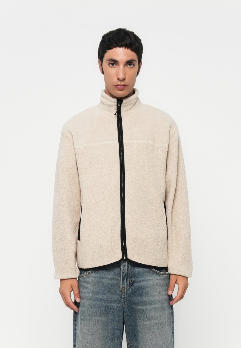 Beige fleecejakke med høy krage, svart glidelås foran og sidelommer, med strukturelt materiale og avslappet passform.
